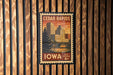 A vintage-style postage stamp from Cedar Rapids, Iowa, featuring a cityscape and a bridge, mounted on a wooden wall.