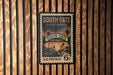 A vintage postage stamp from the United States, featuring a street scene with palm trees and buildings, mounted on a wooden wall.