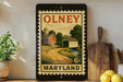 A framed vintage-style stamp depicting a rural scene in Maryland, with a barn, field, and stream. The stamp is placed on a shelf next to a wooden cutting board and a potted plant.