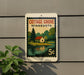 A vintage Minnesota postage stamp featuring a cottage and a lake, mounted on a wall next to a plant.