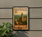 A vintage-style postage stamp from Arkansas featuring a building and landscape, mounted on a wall next to a plant.