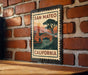 A metal sign with a vintage-style illustration of a bridge and a tree, mounted on a brick wall.