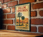 A vintage-style metal sign with the text "PALM SPRINGS" and "8¢ CALIFORNIA" printed on it, mounted on a brick wall.