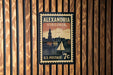 A vintage-style postage stamp from the United States, featuring the city of Alexandria, Virginia, with a sailboat and a building in the background.