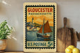 A vintage postage stamp from Gloucester, Massachusetts, featuring a sailboat on the water, is displayed on a shelf next to a wooden cutting board and some lemons.