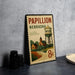 A vintage-style metal sign with the text "PAPILLON NEBRASKA" and an illustration of a water tower, placed on a gray countertop next to some black camera lenses.