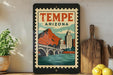 A framed poster of Tempe, Arizona, featuring a bridge and mountains, is displayed on a shelf next to a wooden cutting board and some lemons.