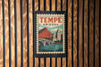A vintage-style poster of Tempe, Arizona, featuring a bridge and mountains, is displayed on a wooden wall.