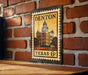 A metal sign with a vintage-style illustration of a building and the text "DENTON TEXAS 6¢" is displayed on a brick wall.