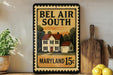 A framed poster of a house with the text "Bel Air South Maryland 15c" on it, placed on a shelf next to a wooden cutting board and a potted plant.