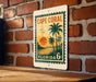 A vintage-style metal sign with a tropical beach scene and palm trees, mounted on a brick wall.