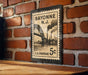 A vintage Bayonne New Jersey postage stamp is displayed on a brick wall, featuring an illustration of a bridge and steam-powered ships.