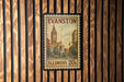 A vintage-style postage stamp from Evanston, Illinois, featuring a church and buildings, is displayed on a wooden wall.