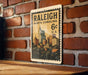 A metal sign with a postage stamp design hangs on a brick wall, featuring the text "RALEIGH NORTH CAROLINA 6¢ U.S."
