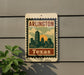 A vintage-style sign with the text "ARLINGTON TEXAS" and a graphic of a city skyline, mounted on a gray wall next to a plant.