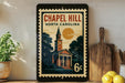 A vintage-style postage stamp depicting a church with a clock tower, surrounded by trees and a large sun, is displayed on a wall next to a wooden cutting board and some lemons.