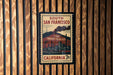 A vintage-style postage stamp from California, featuring an illustration of a city skyline and industrial buildings, mounted on a wooden wall.