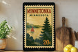 A framed vintage-style postage stamp depicting Minnetonka, Minnesota, with a sailboat on a lake and a pine tree in the background.
