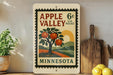 A vintage-style postage stamp from Minnesota, featuring an illustration of an apple tree with oranges, is displayed on a shelf next to a wooden cutting board and a potted plant.