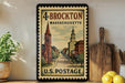 A framed vintage postage stamp from Massachusetts, featuring a street scene with a church and buildings, is displayed on a shelf next to a wooden cutting board and some lemons.