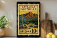 A framed poster of Catalina Foothills, a desert landscape with mountains in the background, is displayed on a wall next to a wooden cutting board and some potted plants.