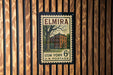A vintage-style postage stamp from the United States, featuring a building and a tree, mounted on a wooden wall.