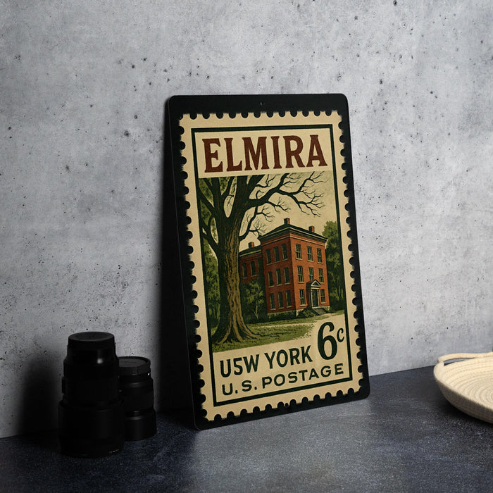 A framed vintage U.S. postage stamp from Elmira, New York, featuring a building and a tree, placed on a countertop next to some black camera lenses.