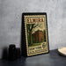 A framed vintage U.S. postage stamp from Elmira, New York, featuring a building and a tree, placed on a countertop next to some black camera lenses.