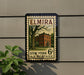 A vintage-style postage stamp featuring a brick building and a tree, mounted on a wall.