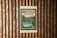 A vintage postage stamp from Prattville, Alabama, featuring a waterfall and buildings, is displayed on a wooden wall.