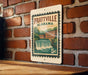 A vintage postage stamp from Prattville, Alabama, featuring a waterfall and buildings, is displayed on a brick wall.