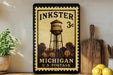 A vintage-style metal stamp depicting a water tower, with the text "INKSTER 3c" and "MICHIGAN U.S. POSTAGE" printed on it. The stamp is displayed on a shelf next to a wooden cutting board and a potted plant.