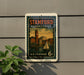 A vintage postage stamp from the United States, featuring a cityscape and a sailboat, is displayed on a wall.