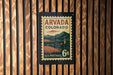 A vintage U.S. postage stamp from Colorado, featuring a mountain landscape and a clock tower, is displayed on a wooden wall.