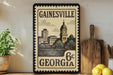 A framed vintage postage stamp from Georgia, featuring a cityscape and a clock tower, is displayed on a shelf next to a wooden cutting board and a potted plant.
