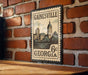 A vintage-style postage stamp from Gainesville, Georgia, featuring a cityscape and a clock tower, mounted on a brick wall.