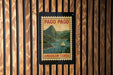 A vintage-style postage stamp depicting a scenic landscape with mountains, a river, and a sailboat, hanging on a wooden wall.