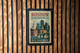 A vintage-style postage stamp from Boston, Massachusetts, featuring a cityscape and a church, mounted on a wooden wall.