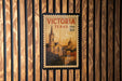 A postage stamp from Victoria, Texas, featuring a church and other buildings, is displayed on a wooden wall.