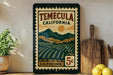 A vintage-style postage stamp from Temecula, California, featuring a landscape with mountains, fields, and a sun. The stamp is displayed on a shelf next to a wooden cutting board and some kitchen utensils.