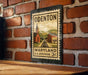 A vintage-style metal stamp depicting a rural scene with a church, train tracks, and a mountain backdrop, mounted on a brick wall.