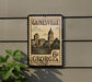 A vintage-style postage stamp from Georgia, featuring a cityscape and a clock tower, is displayed on a wall.