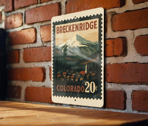 A metal sign with a mountain landscape and a small town in the background, mounted on a brick wall.