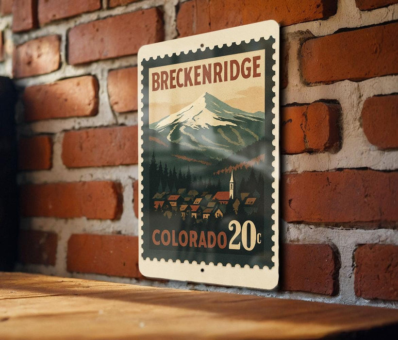A metal sign with a mountain landscape and a small town in the background, mounted on a brick wall.