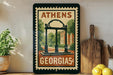 A framed poster of the University of Georgia's Arch, a historic building, is displayed on a shelf next to a wooden cutting board and some lemons.