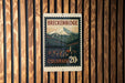 A vintage-style metal sign with a mountain landscape and a small town in the background, mounted on a wooden wall.