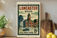 A vintage-style postage stamp from the United States Postal Service, featuring an illustration of a town with a clock tower, is displayed on a shelf next to a wooden cutting board and some potted plants.