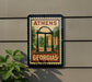 A framed poster of the University of Georgia's iconic arch, displayed on a wall next to a plant.