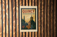 A vintage-style poster of QuINCY, Massachusetts, featuring a church and other buildings, is displayed on a wooden wall.