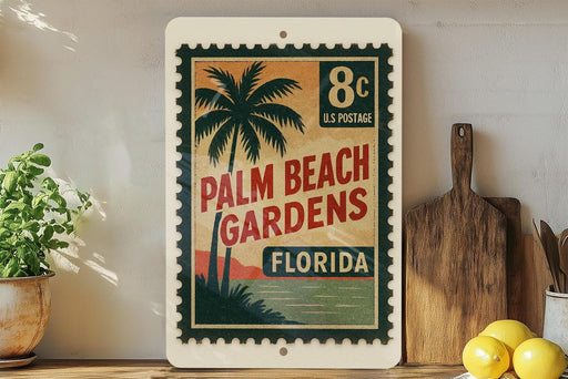 A vintage-style postage stamp featuring palm trees and a beach scene, with the text "Palm Beach Gardens, Florida" printed on it. The stamp is displayed on a shelf next to a wooden cutting board and some kitchen utensils.